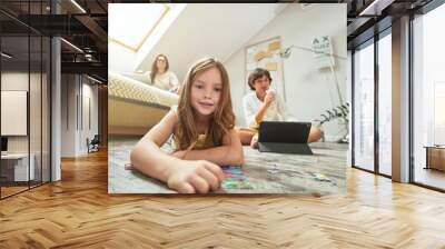 Family staying at home. Cute little girl lying on the floor and smiling at camera while playing with puzzles, spending time together with brother and mother in the living room Wall mural