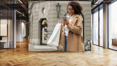 Fashion afro american woman after shopping with paper bags standing in city and using phone Wall mural