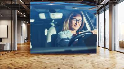 Happy attractive caucasian woman wearing eyeglasses driving her modern car through the city on sunny day, she is looking on the road and smiling Wall mural