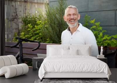 Happy guy. Successful modern middle aged businessman in stylish suit smiling aside while working on his laptop, sitting on the bench outdoors with a bicycle next to him Wall mural