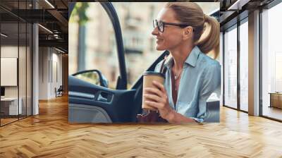 Happy successful middle aged business woman holding paper cup of coffee while getting out of her car, she is arriving at work Wall mural