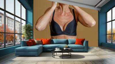 Relaxed mature woman with fit body in black underwear touching her head and smiling with eyes closed while standing and posing half naked isolated over beige background Wall mural