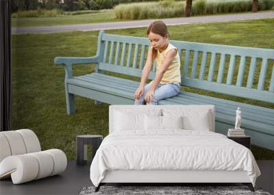 Sad little girl sitting on the bench in park on a warm day, waiting for parents outdoors Wall mural