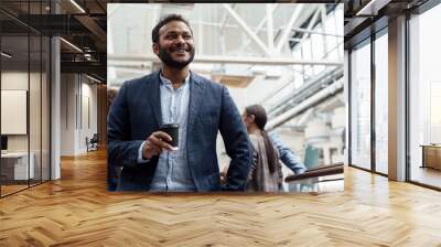 Smiling indian businessman drinking coffee and looking side while standing in office Wall mural