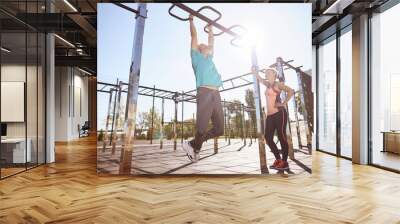 Strong senior man in sportswear doing pull-ups on horizontal bar while training with his wife outdoors. Full length. Beautiful aged couple exercising in the morning Wall mural