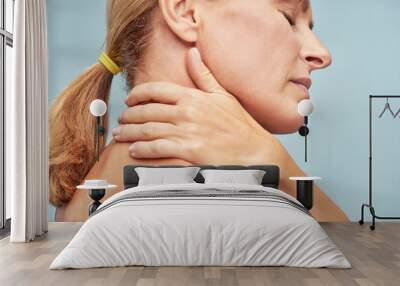 Suffering from back pain. Rear view of a mature woman touching her neck while standing against blue background Wall mural