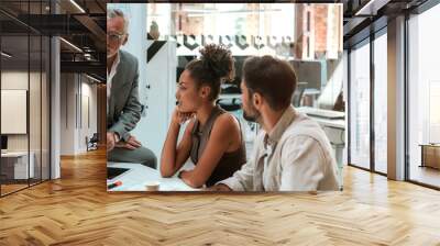 Working process. Mature businessman talking about something with his young colleagues while sitting together at the office table in the modern office Wall mural