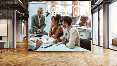 Working process. Mature businessman talking about something with his young colleagues while sitting together at the office table in the modern office Wall mural