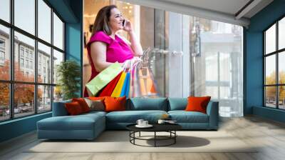 Laughing glamour plus size lady talking smartphone with paper shopping bag at modern boutique mall Wall mural