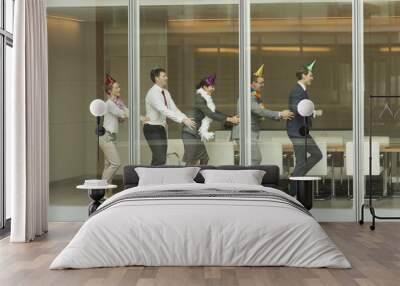 Business people wearing party hats dancing in conga line at conference room window Wall mural