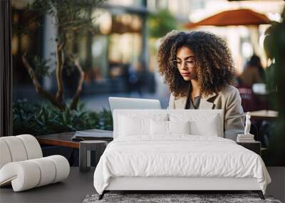 A woman with curly hair is sitting at a table with a laptop in front of her Wall mural