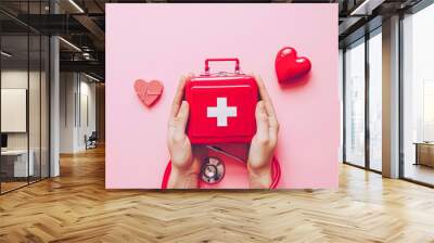 Hands presenting a first aid kit with a red stethoscope and heart shapes on a pink background, symbolizing healthcare. Wall mural