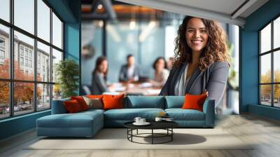 Young smiling successful businesswoman in formal wear standing in boardroom with arms crossed and looking at camera.In background her colleagues having meeting. Wall mural