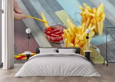 Hand dipping French fries on decorative fork in ketchup. Hand dipping French fries in ketchup. French fries serve in two glass mugs on a wooden surface painted in different colors. Wall mural