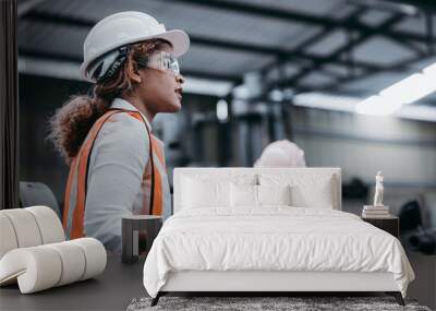 Female industrial engineer wearing a white helmet while standing in a heavy industrial factory behind she talking with workers, Various metal parts of the project Wall mural