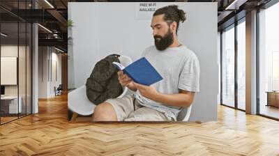 a bearded young man reads a blue book while seated in a bright indoor waiting room with a backpack a Wall mural