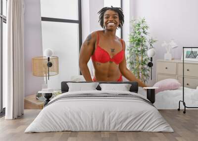 A cheerful african american woman in red lingerie stands confidently in a bright bedroom setting, embodying positivity and comfort. Wall mural