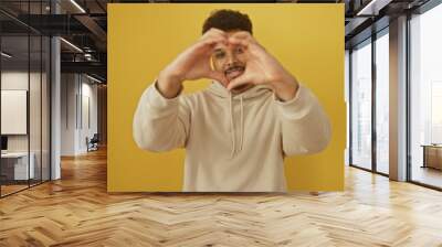 A cheerful african man in casual attire making a heart shape with his hands against a vibrant yellow background. Wall mural