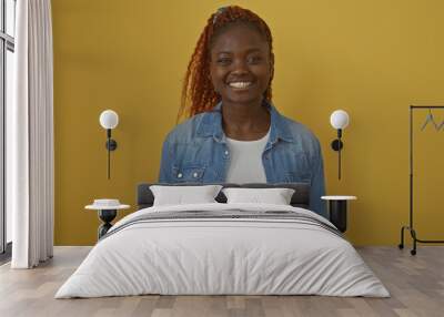 A cheerful young african woman in casual denim attire posing against a vibrant yellow background with a genuine smile. Wall mural