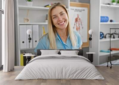 A cheerful young blonde woman medical professional standing in a clinic room with shelves and anatomy posters. Wall mural