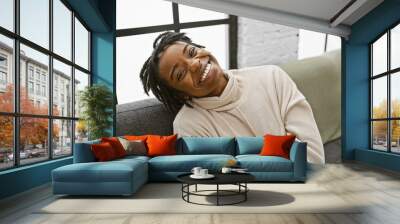 A cheerful young woman with dreadlocks lounging on a sofa indoors smiling at the camera. Wall mural