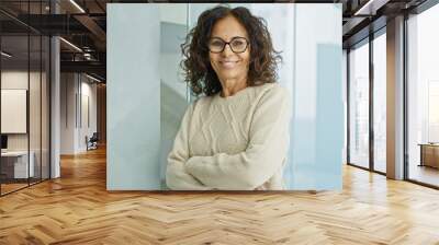 A confident middle-aged hispanic woman with arms crossed poses in a modern office interior. Wall mural