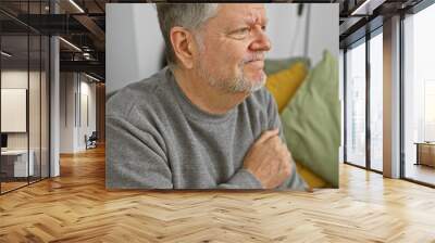 A contemplative senior man sits indoors on a couch, with a neutral expression and casual attire against a cozy home background. Wall mural