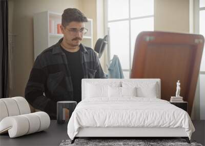 A contemplative young hispanic man with a moustache and glasses stands in a well-lit dressing room, selecting clothes. Wall mural