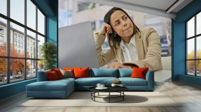 A contemplative young hispanic woman in business casual attire sits by a laptop at a cafe with a blurred background. Wall mural