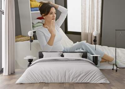 A contemplative young hispanic woman sits on a bed in a bedroom, displaying a casual and relaxed posture. Wall mural