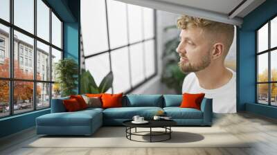 A contemplative young man with blue eyes and a beard in casual attire inside a modern room with a large window and lush plant decor. Wall mural