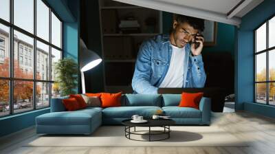 A focused hispanic man in casual attire talks on the phone while working late in an office illuminated by a desk lamp. Wall mural