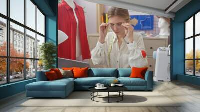 A focused woman tailor adjusts her glasses in a colorful fashion atelier surrounded by sewing equipment and mannequins. Wall mural