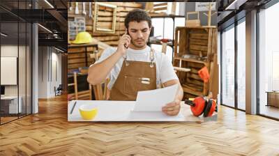 A focused young man in a workshop talks on the phone while examining a document, surrounded by woodwork tools. Wall mural