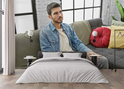 A handsome young hispanic man in casual attire lounges thoughtfully on a sofa in a stylish modern living room. Wall mural