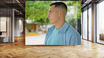 A handsome young hispanic man in casual attire stands thoughtfully in a lush outdoor park setting. Wall mural