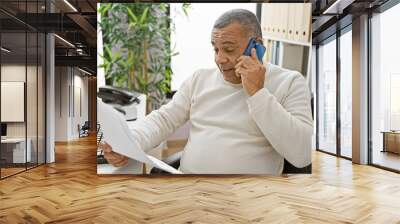 A mature hispanic man talks on a smartphone, reviewing paperwork in a modern office with plants. Wall mural