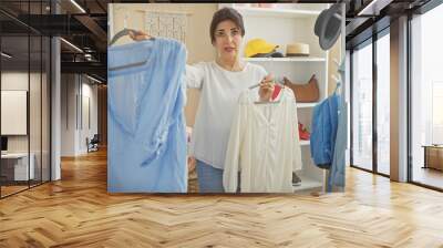 A middle-aged hispanic woman compares blouses in a bright dressing room, surrounded by clothes and accessories. Wall mural