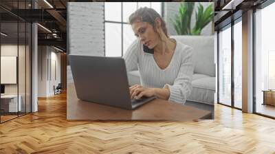 A multitasking young woman works on her laptop with a phone held between shoulder and ear in a bright home interior. Wall mural