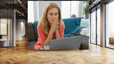 A pensive middle-aged woman sits at home with a laptop, showing a moment of concern or decision-making in a cozy living room. Wall mural