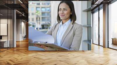 A professional hispanic woman in a beige suit extends documents outdoors in an urban setting. Wall mural