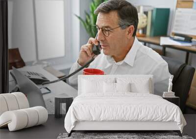 A professional middle-aged man is engaged in a conversation on the phone in a modern office setting, holding a red mug. Wall mural