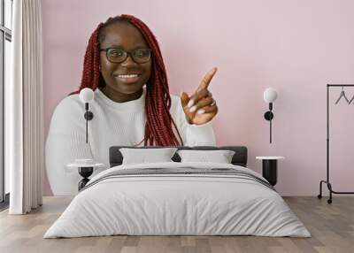 A smiling african american woman with braids wearing glasses and a white sweater poses against a pink background, pointing with her finger. Wall mural