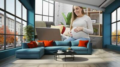 A smiling pregnant hispanic woman sits comfortably at home with a laptop, exuding maternal warmth in a cozy living room. Wall mural