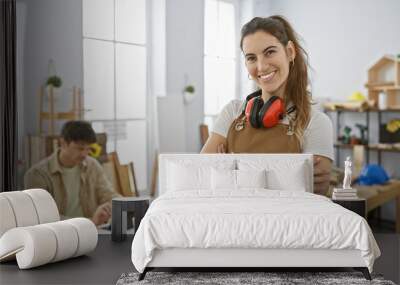 A smiling woman in an apron with headphones and a focused man woodworking in a bright carpentry workshop. Wall mural