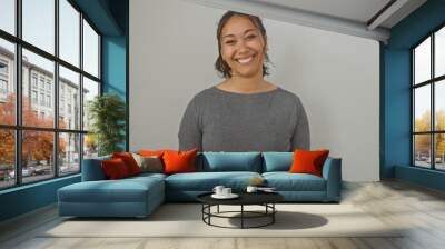 A smiling young adult hispanic woman poses in casual clothing against an isolated white wall background. Wall mural