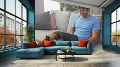 A smiling young man in casual attire works on his laptop while sitting on a gray sofa next to his resting siamese cat in a cozy living room. Wall mural