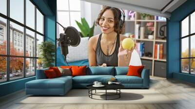 A smiling young woman with headphones holding an apple in a modern radio studio, representing health and lifestyle podcasting. Wall mural