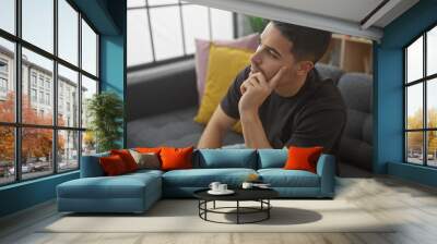 A thoughtful hispanic man with a beard sitting in a modern living room, portraying casual relaxation at home. Wall mural