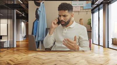 A young, bearded hispanic man wearing a white shirt looks concerned while talking on a smartphone at home, by a desk with a laptop. Wall mural